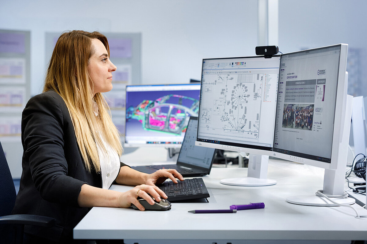 Person an einem Schreibtisch mit technischen Zeichnungen auf den Bildschirmen.