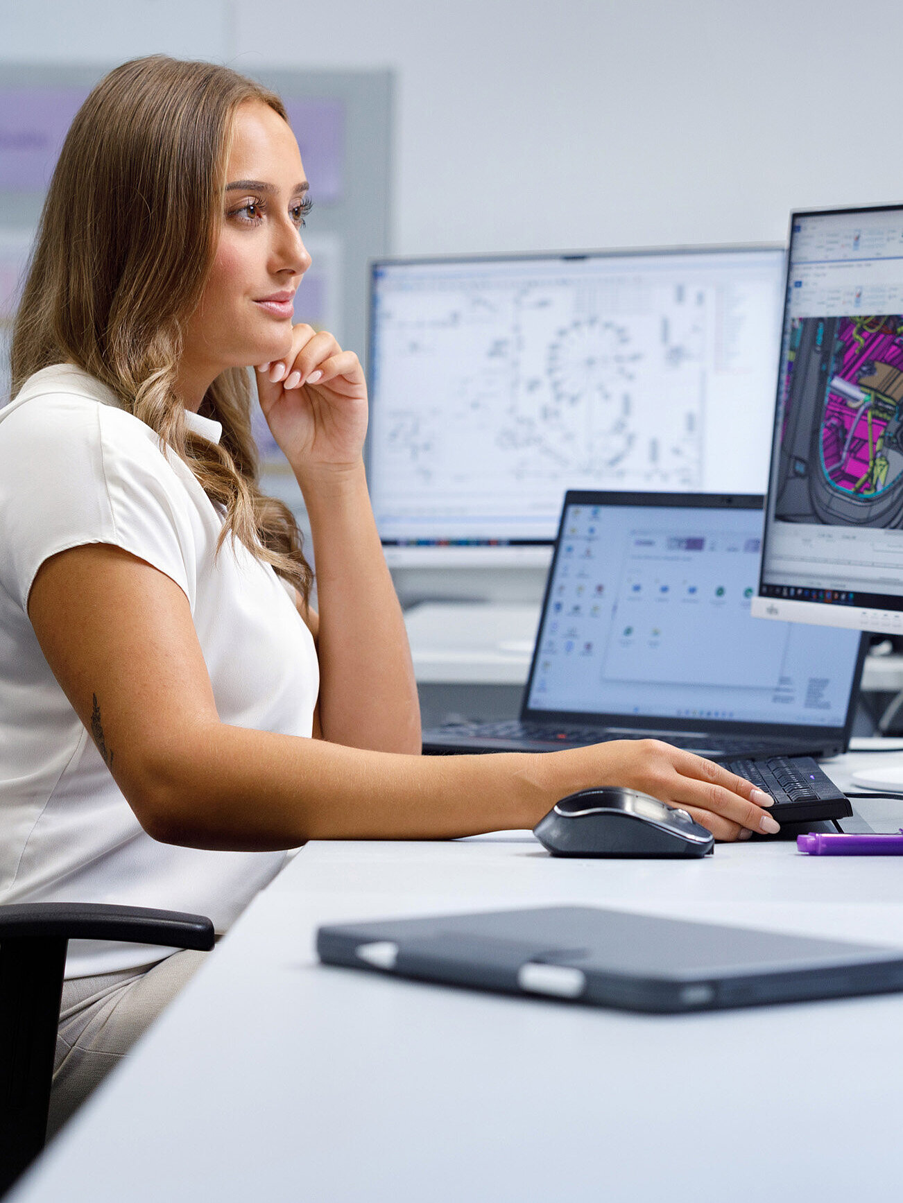 Person an einem Schreibtisch mit technischen Zeichnungen auf den Bildschirmen.